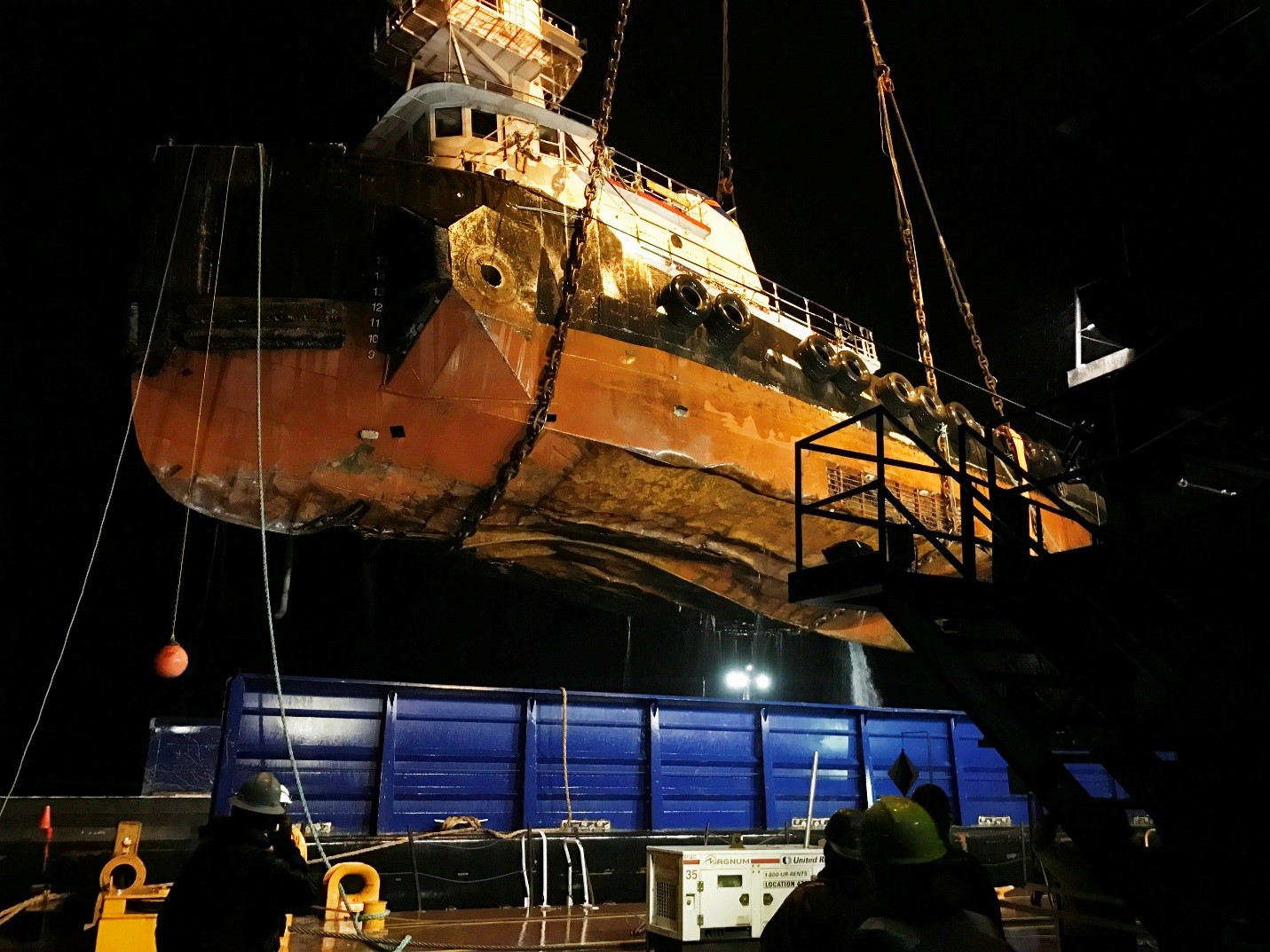 A ship is seen from below being hoisted above the water. This photograph was taken at night.
